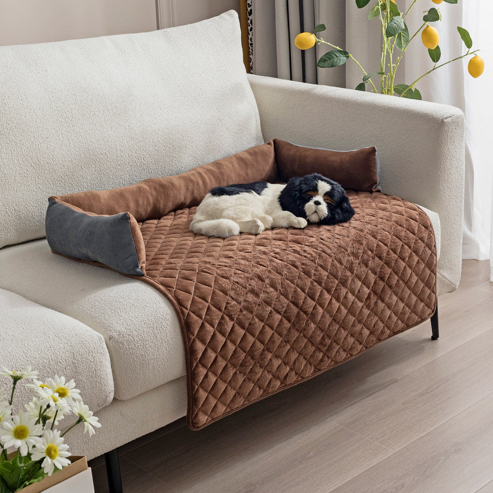 Dog lying on a brown quilted pet bed on a sofa with decorative pillows and flowers.